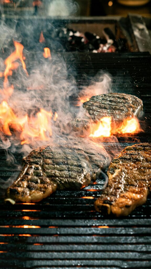 pexels-photo-36543125-36543125 Three flavorful steaks grilling over open flames with smoke rising, creating a savory barbecue scene.